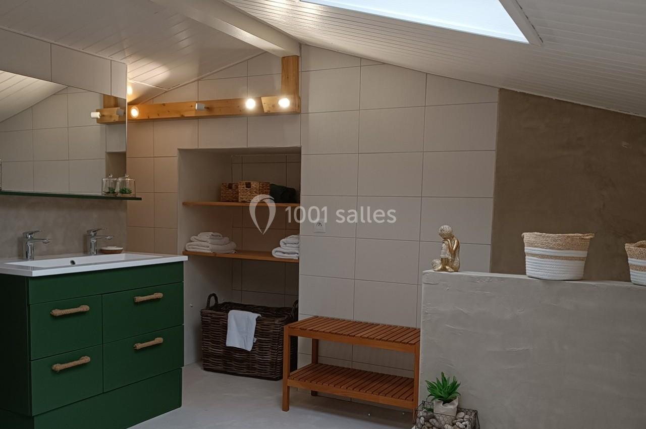 Salle de bain sous combles avec meuble vert, étagères en bois, paniers tressés et lumière naturelle d'un velux.