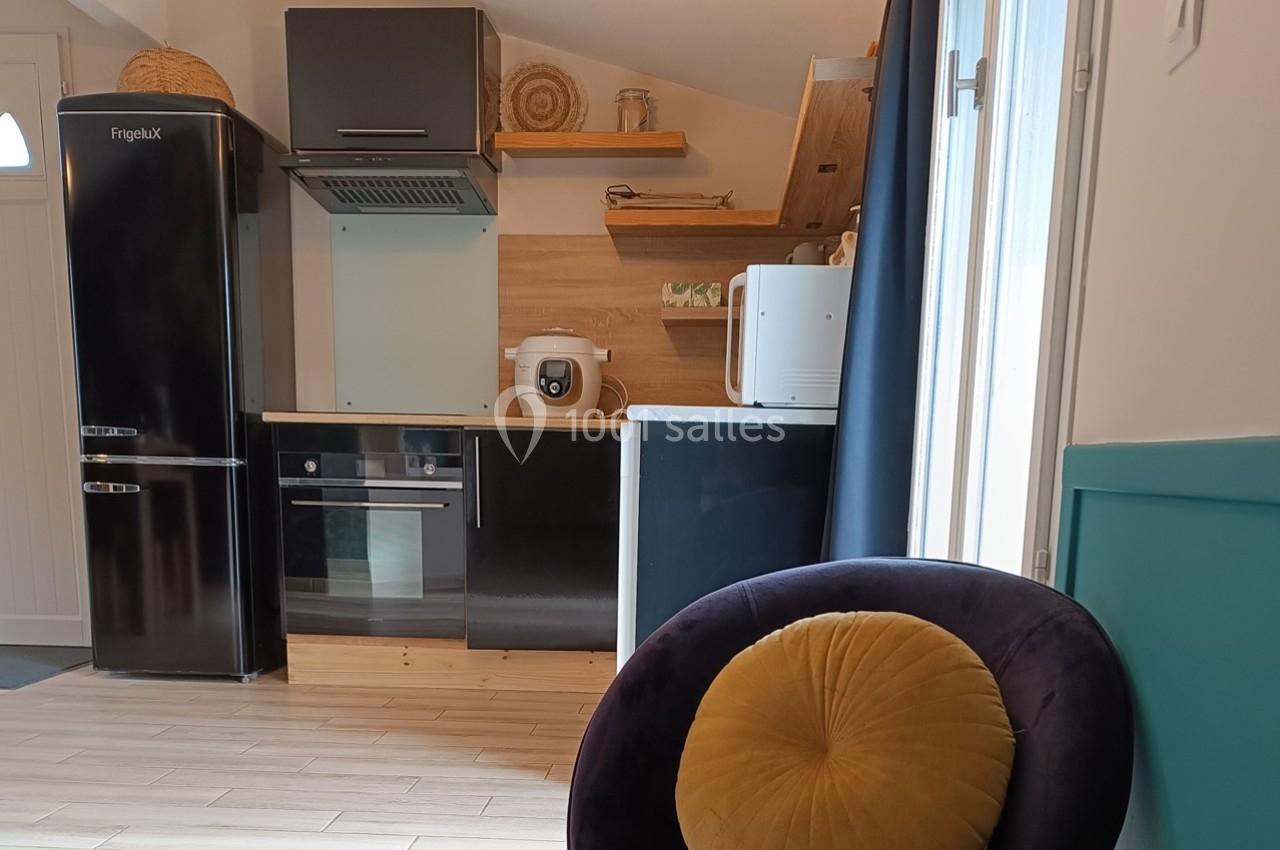 Cuisine moderne avec réfrigérateur noir, four encastré, micro-ondes, étagères en bois et chaise avec coussin jaune.