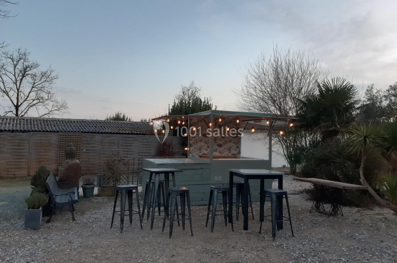 Kiosque extérieur en bois avec tabourets et tables hautes, éclairé par des guirlandes lumineuses, entouré de végétation.