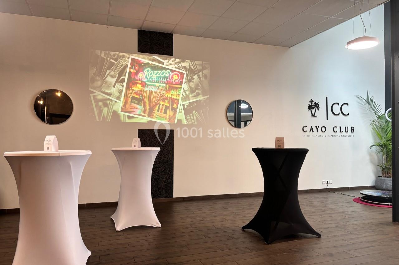 Salle avec tables hautes drapées, projecteur affichant une image, murs décorés de miroirs et logo ’Cayo Club’.