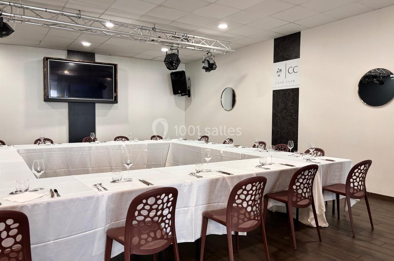 Salle de réunion avec tables disposées en U, nappes blanches, chaises marron et écran mural.