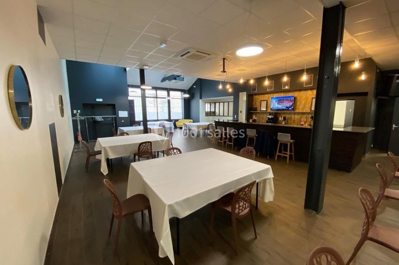 Salle lumineuse avec tables dressées, chaises, bar éclairé et décor moderne, vue sur des baies vitrées.