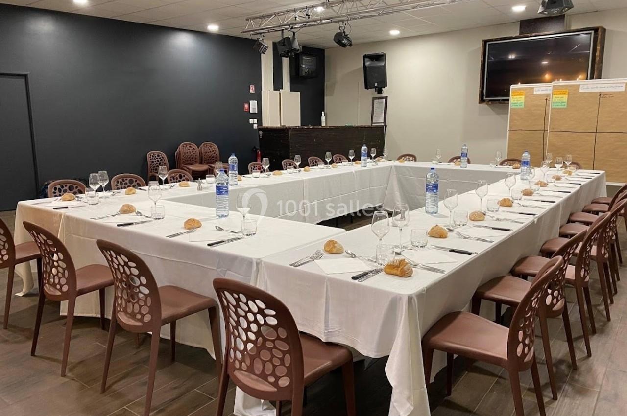 Salle de réception avec tables en U dressées, nappes blanches, couverts, verres, bouteilles d'eau et petits pains.
