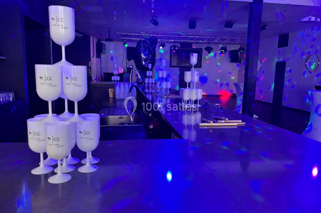 Salle de réception avec un bar éclairé par des lumières colorées et des verres empilés au premier plan.