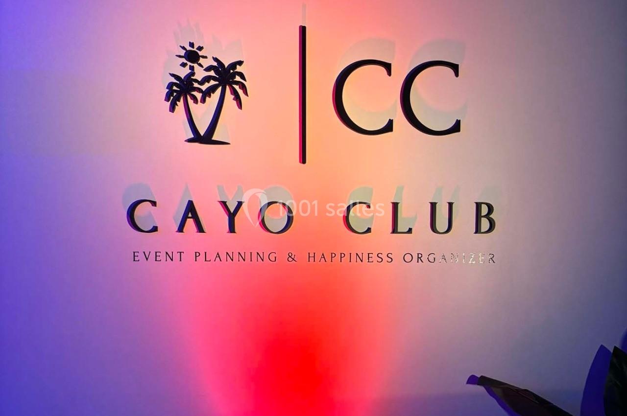 Logo du Cayo Club avec des palmiers, éclairé par des lumières colorées rouge et bleue en arrière-plan.
