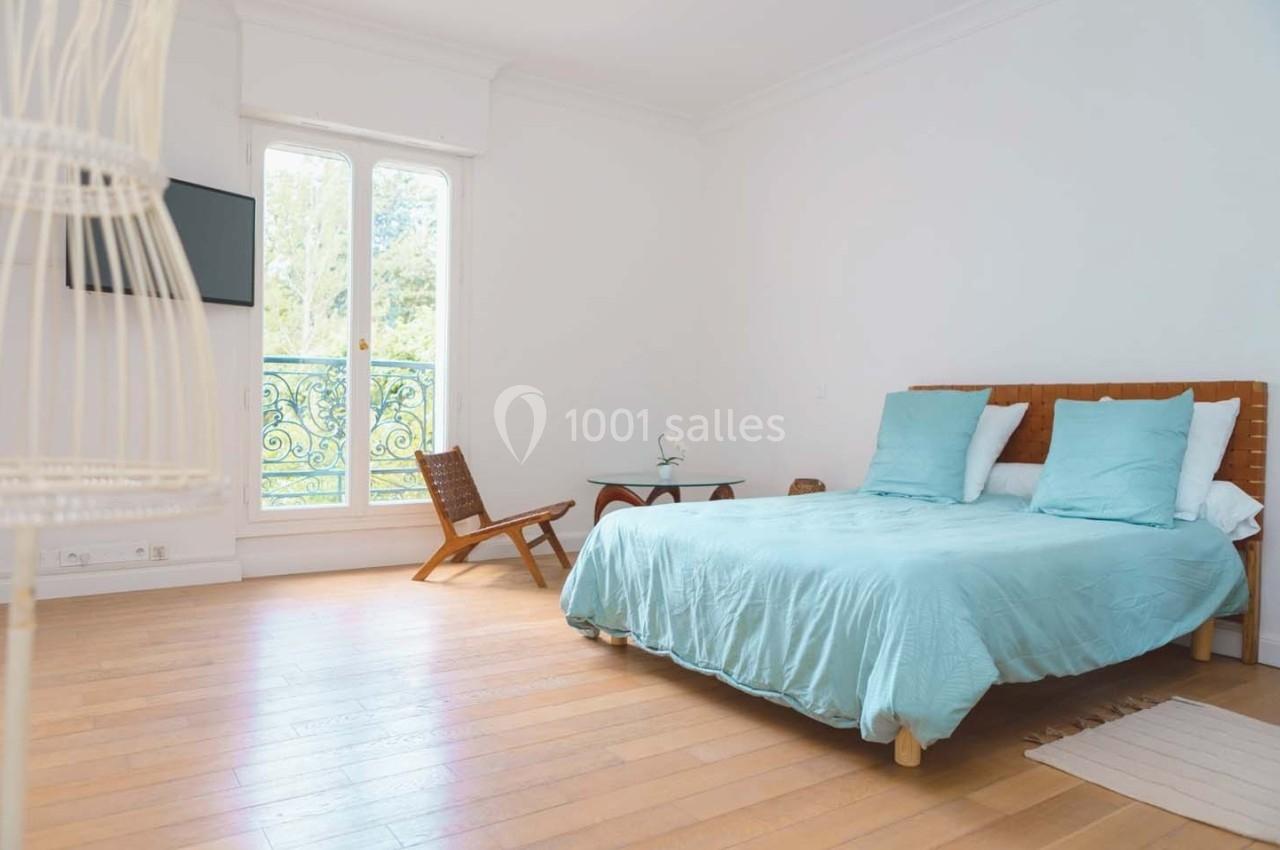 Chambre lumineuse avec lit double, draps bleus, chaise en bois, table basse et grande fenêtre donnant sur un balcon.