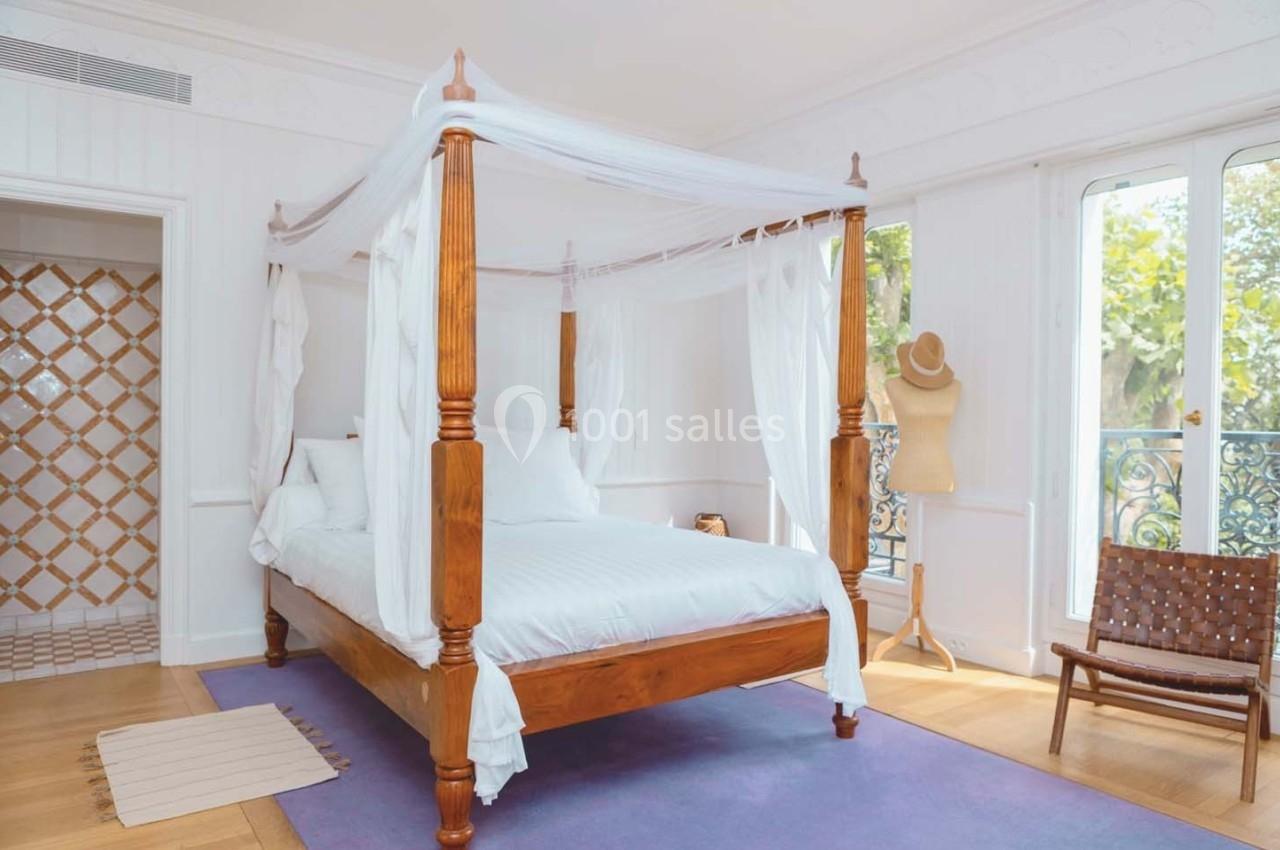 Lit à baldaquin en bois avec voilages blancs dans une chambre lumineuse, parquet clair et vue sur un balcon.