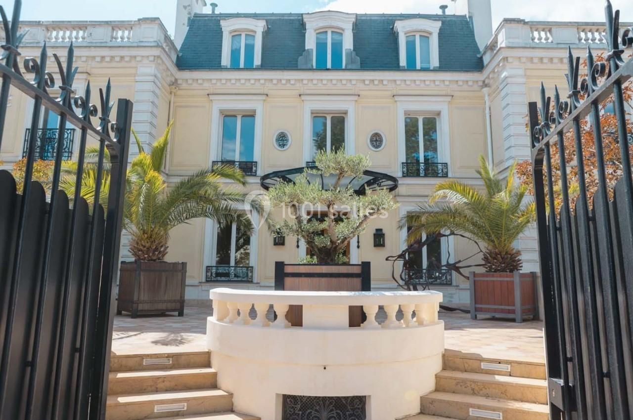 Façade d'un bâtiment élégant de style classique avec des palmiers et une entrée encadrée par des grilles noires.