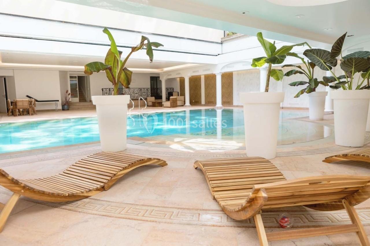 Piscine intérieure entourée de transats en bois et de grandes plantes en pots blancs dans un espace lumineux.