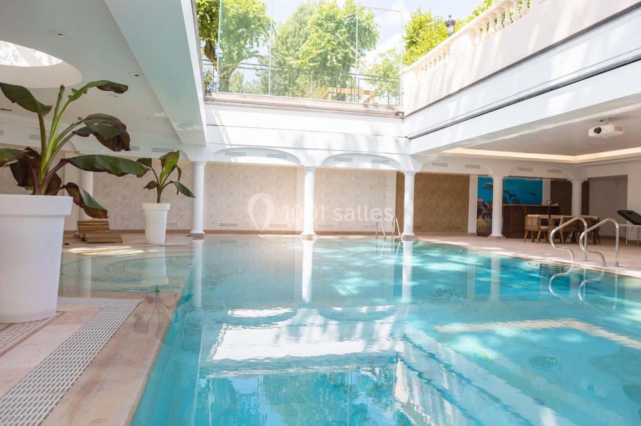 Piscine intérieure lumineuse avec baies vitrées, plantes en pot et mobilier en arrière-plan.