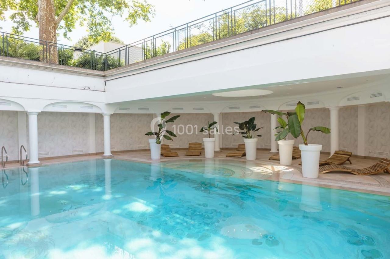 Piscine intérieure lumineuse entourée de transats en bois et de plantes en pots, avec plafond ouvert partiellement.