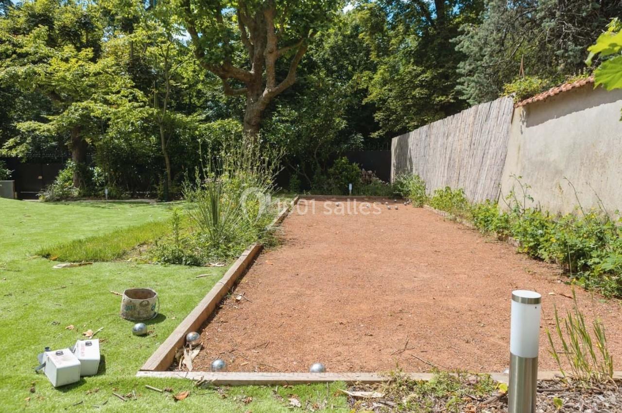 Terrain de pétanque en gravier entouré de végétation, situé dans un jardin avec pelouse et arbres.