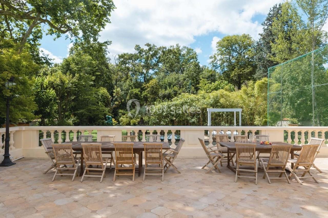 Grande terrasse avec deux longues tables en bois entourées de chaises, donnant sur un jardin verdoyant et arboré.