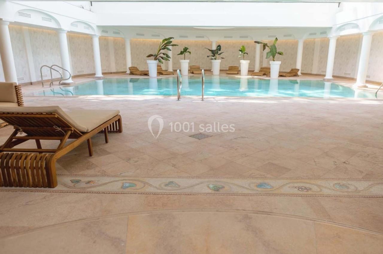 Piscine intérieure entourée de transats en bois et de grandes plantes en pot, dans un espace lumineux et élégant.