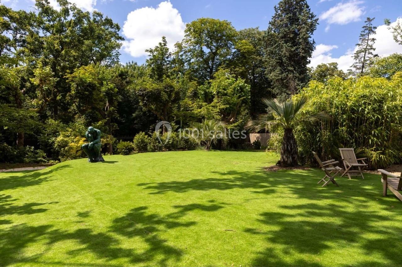 Pelouse verdoyante entourée d'arbres et de bambous, avec une statue et des chaises en bois sous un ciel ensoleillé.