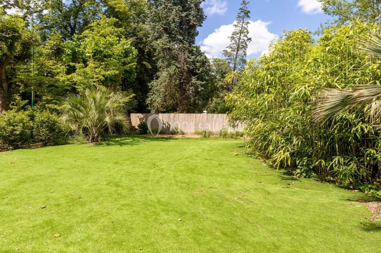 Pelouse verte entourée de végétation dense, avec des arbres et des bambous sous un ciel partiellement nuageux.