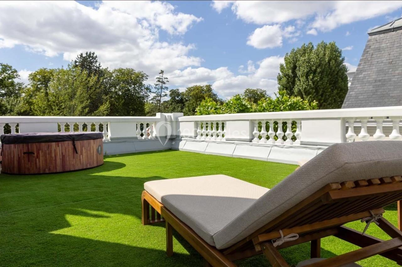 Terrasse avec pelouse artificielle, bain à remous en bois et chaise longue sous un ciel partiellement nuageux.