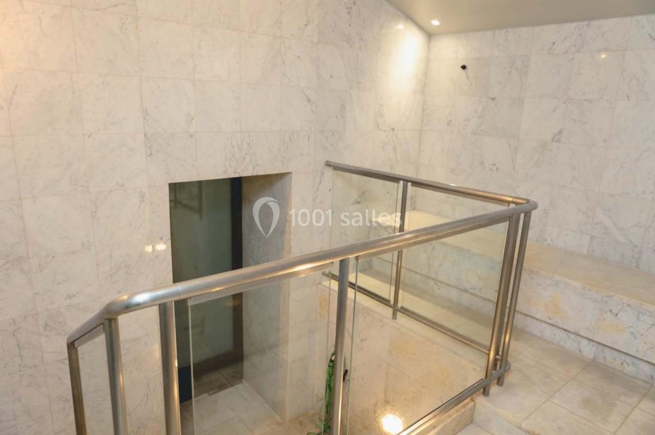 Escalier en marbre blanc avec rambarde en métal et parois vitrées, éclairé par des spots au plafond.