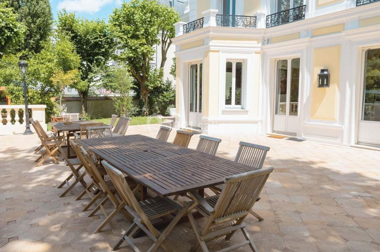 Grande terrasse pavée avec deux longues tables en bois et des chaises pliantes, entourée d'arbres et d'une façade claire.