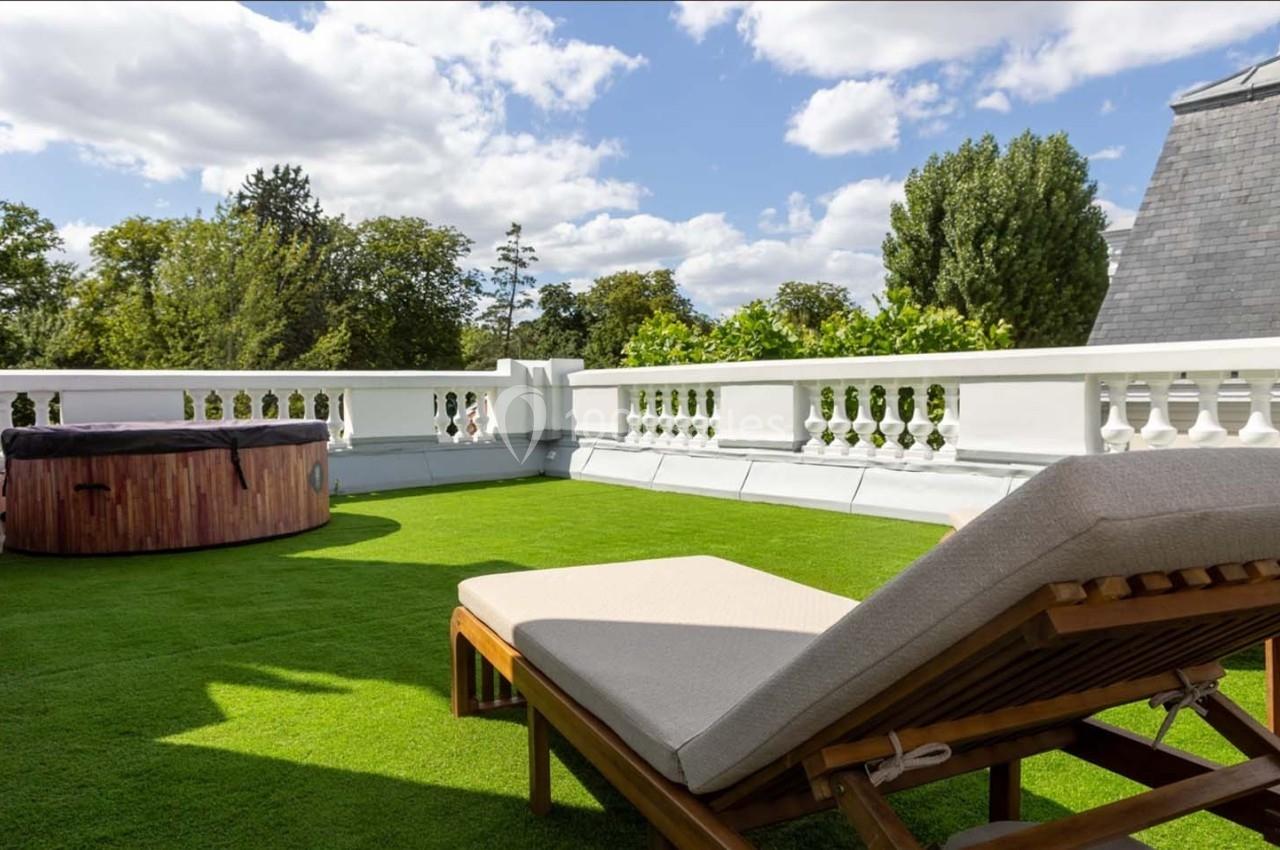 Terrasse avec pelouse artificielle, bain à remous en bois et transat, entourée de balustrades blanches sous un ciel dégagé.