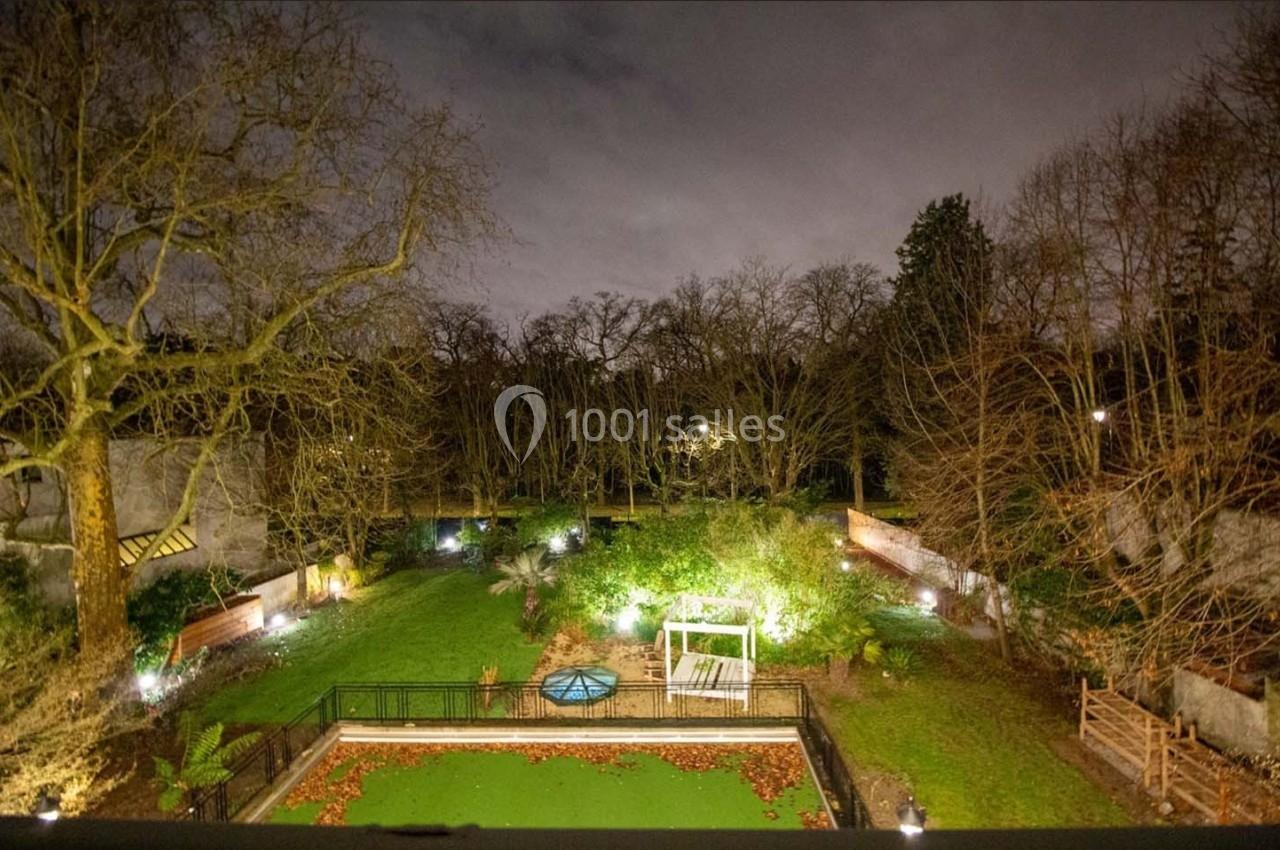 Vue nocturne d'un jardin éclairé avec pelouse, arbres dénudés, bassin central et clôtures en arrière-plan.