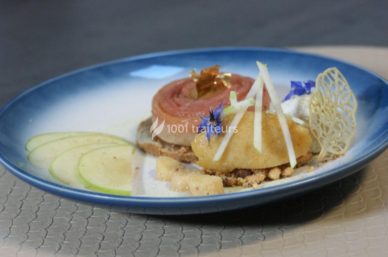 Dessert raffiné composé de pommes, crumble, fleurs comestibles et décorations, présenté sur une assiette bleue.