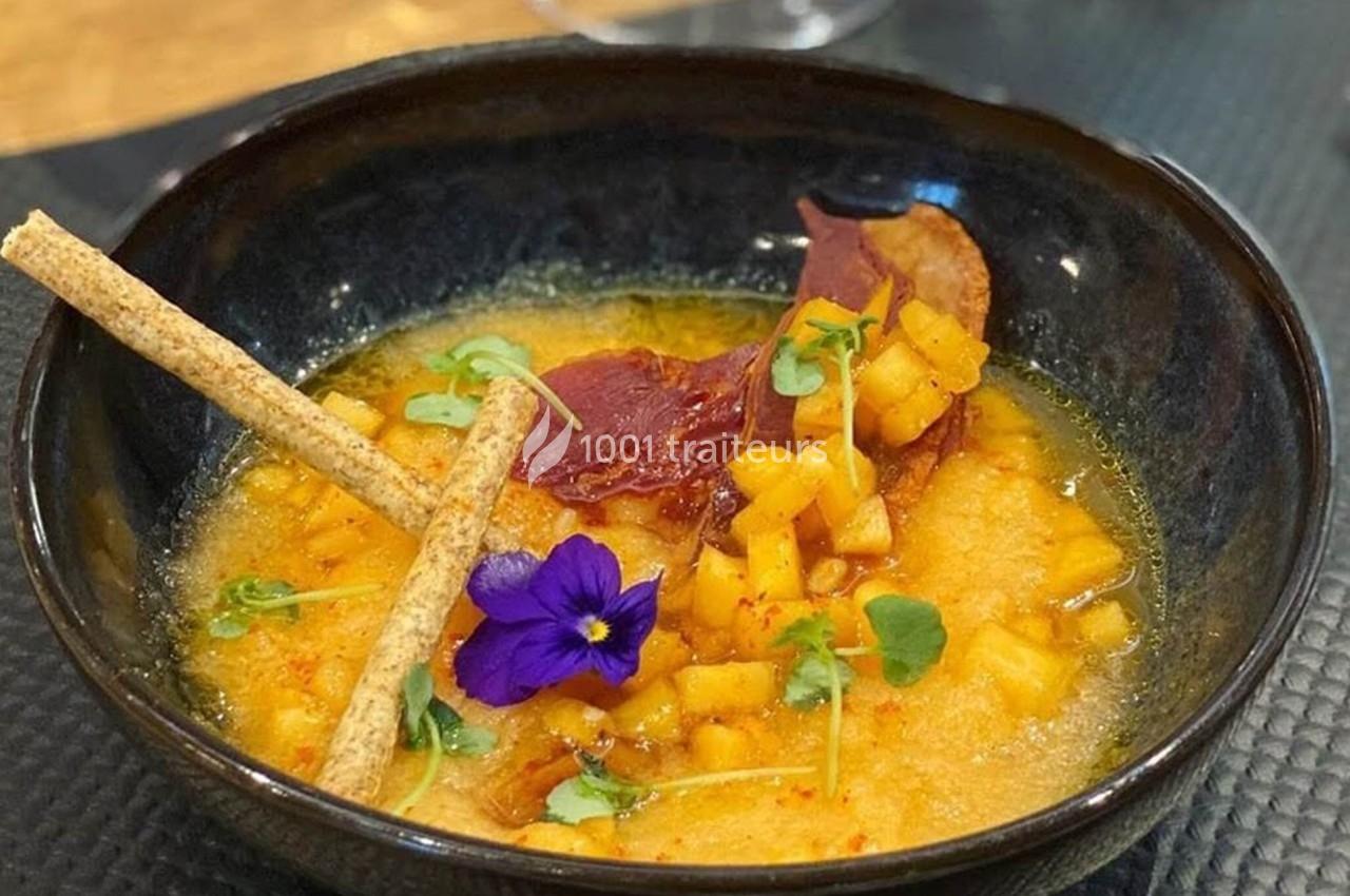 Soupe garnie de dés de légumes, chips de jambon, fleurs comestibles et gressins dans un bol noir.