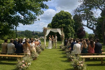 Un couple échange ses vœux sous une arche fleurie lors d'une cérémonie de mariage en plein air, entouré d'invités.
