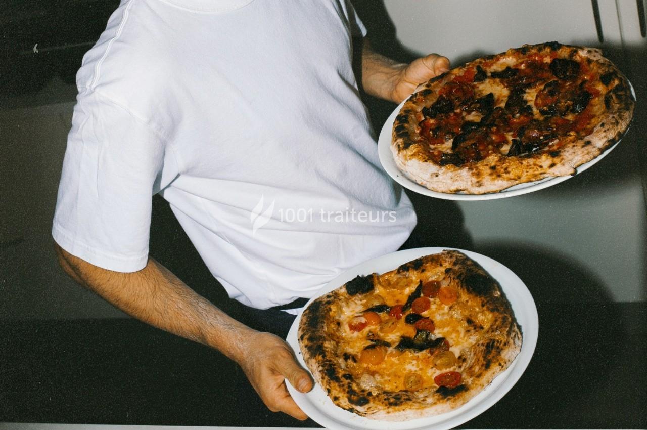 Un homme en t-shirt blanc tenant deux pizzas garnies de légumes et de charcuterie sur des assiettes blanches.