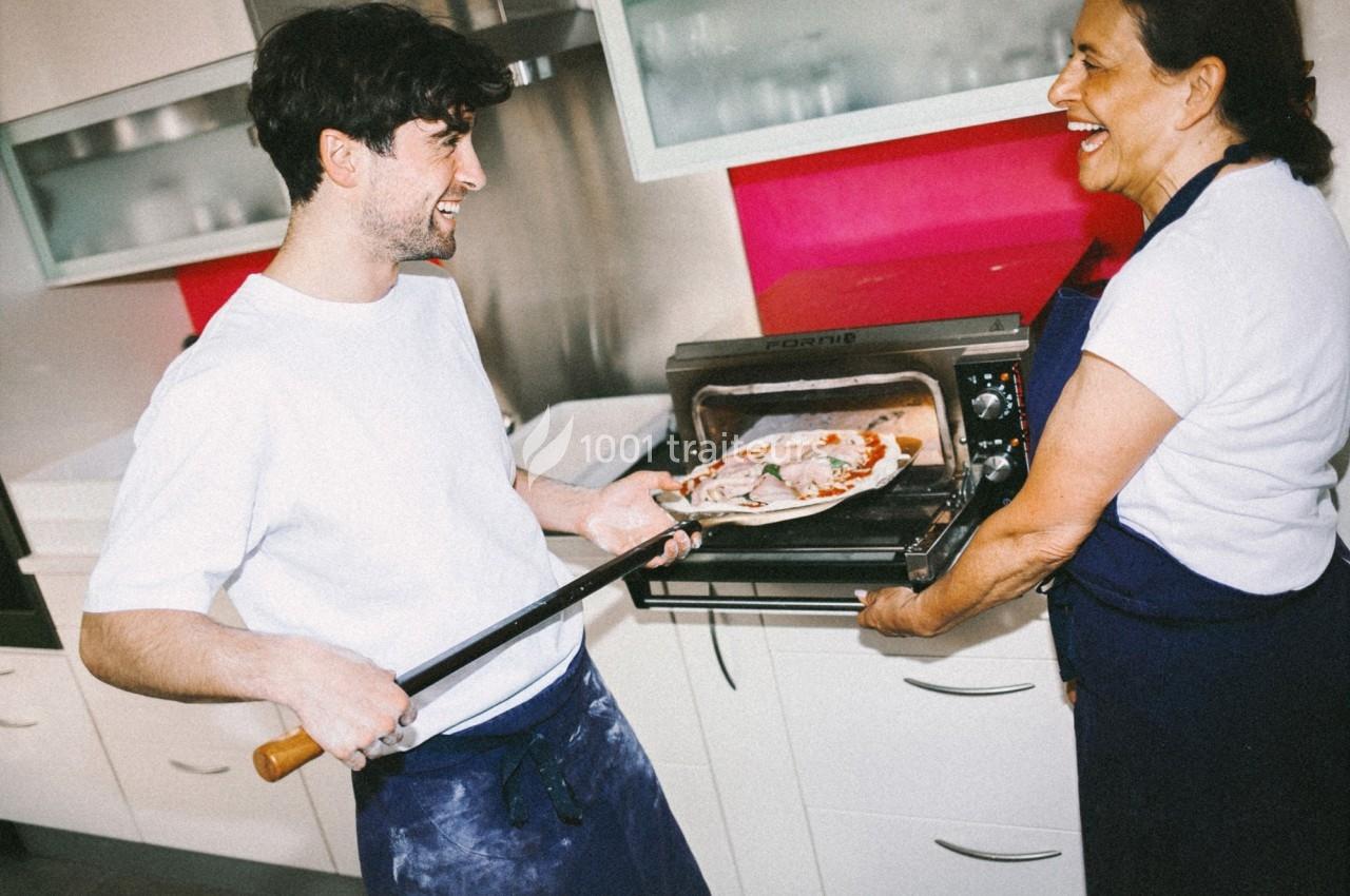 Un homme retire une pizza du four à l'aide d'une pelle, accompagné d'une femme souriante dans une cuisine moderne.