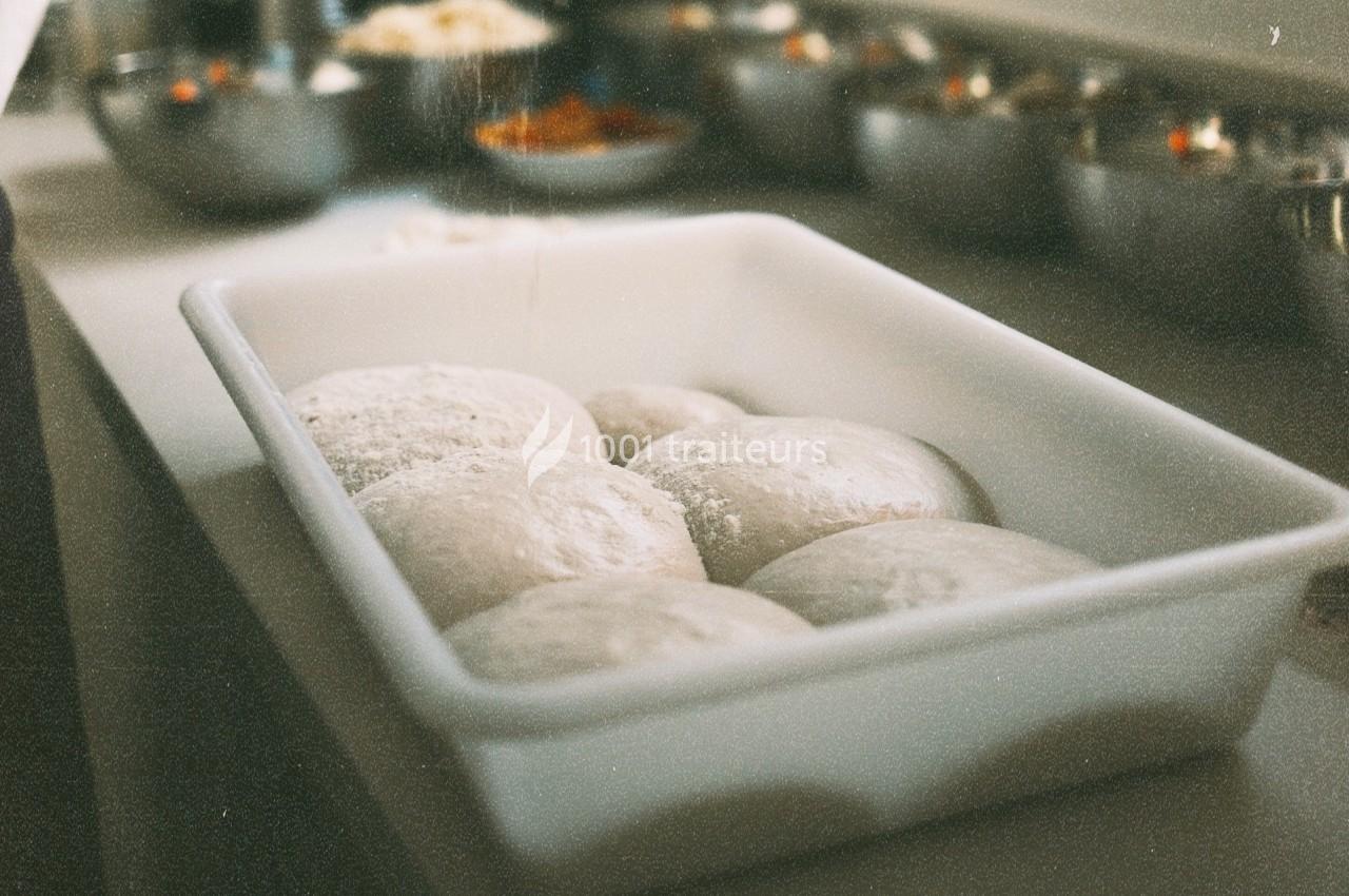Pâte levée en boule dans un plat rectangulaire blanc, avec des bols d'ingrédients flous en arrière-plan.