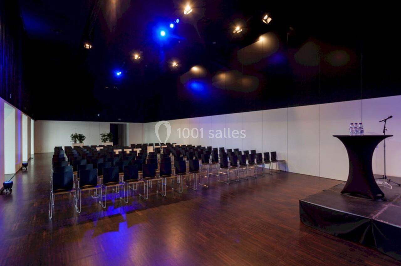 Salle de conférence vide avec rangées de chaises noires, éclairage bleu et une table haute sur une estrade.