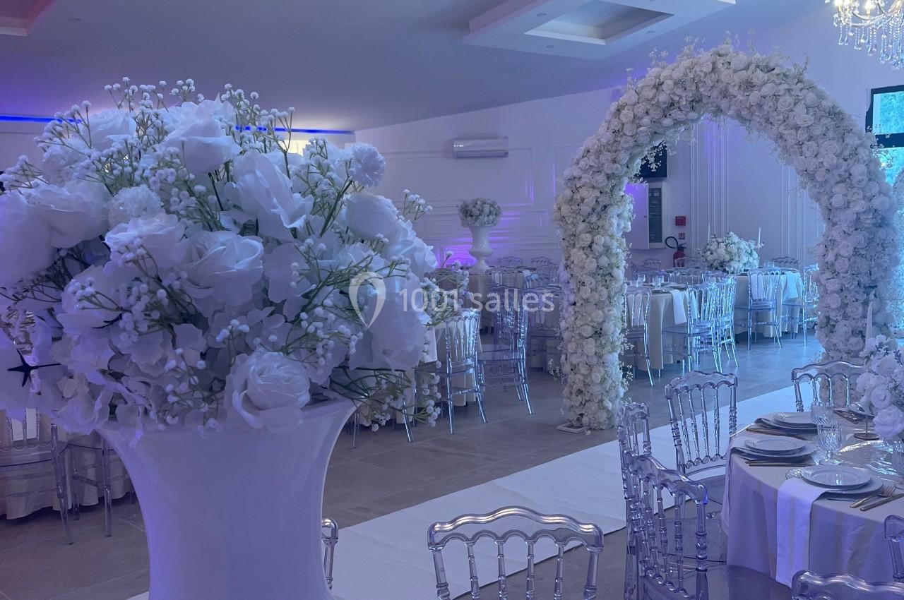 Salle de réception décorée avec des fleurs blanches, des tables dressées et une arche florale illuminée.