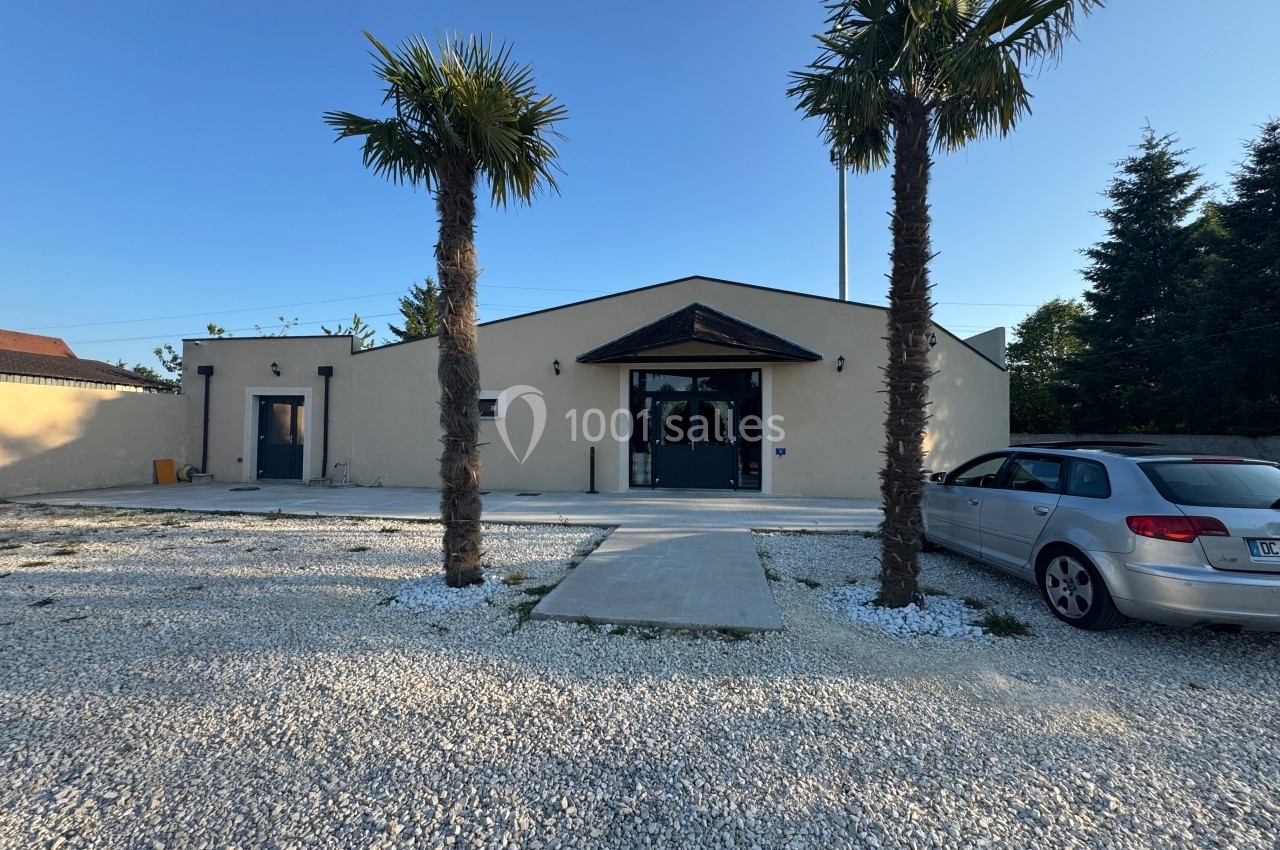 Façade d'un bâtiment beige avec deux palmiers devant, une voiture stationnée à droite et un sol en gravier.