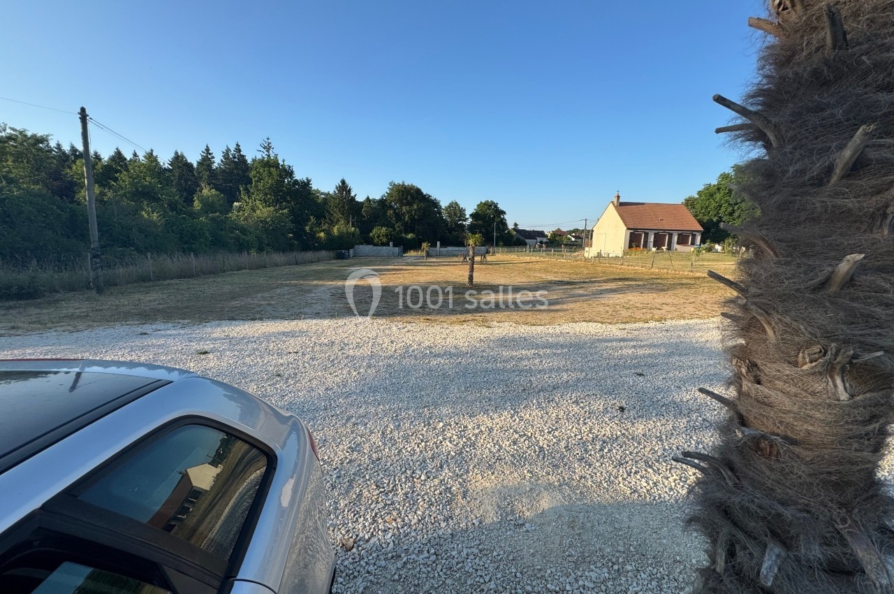 Vue d'un terrain dégagé avec gravier, pelouse, une maison au fond et un arbre au premier plan à droite.
