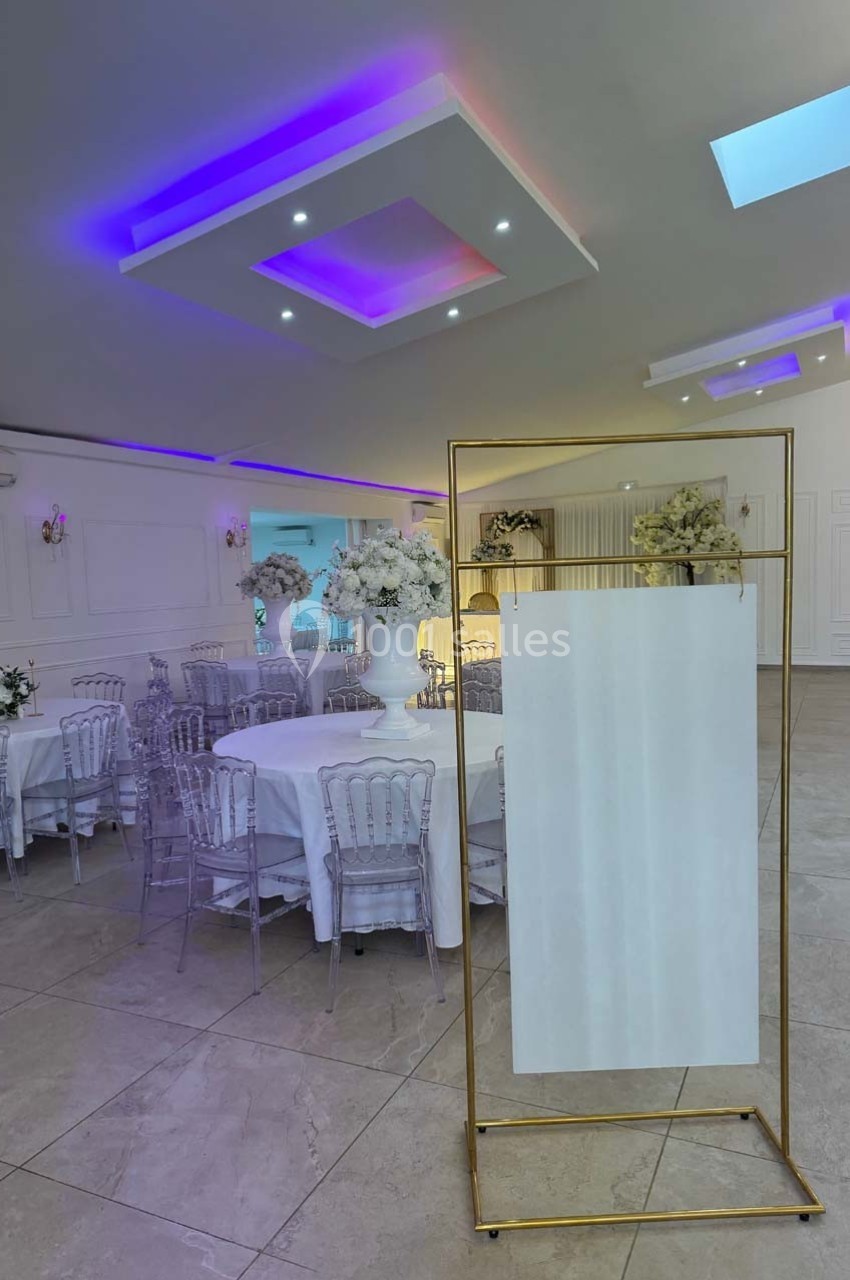 Salle de réception élégante avec tables rondes dressées, chaises transparentes et décorations florales blanches.