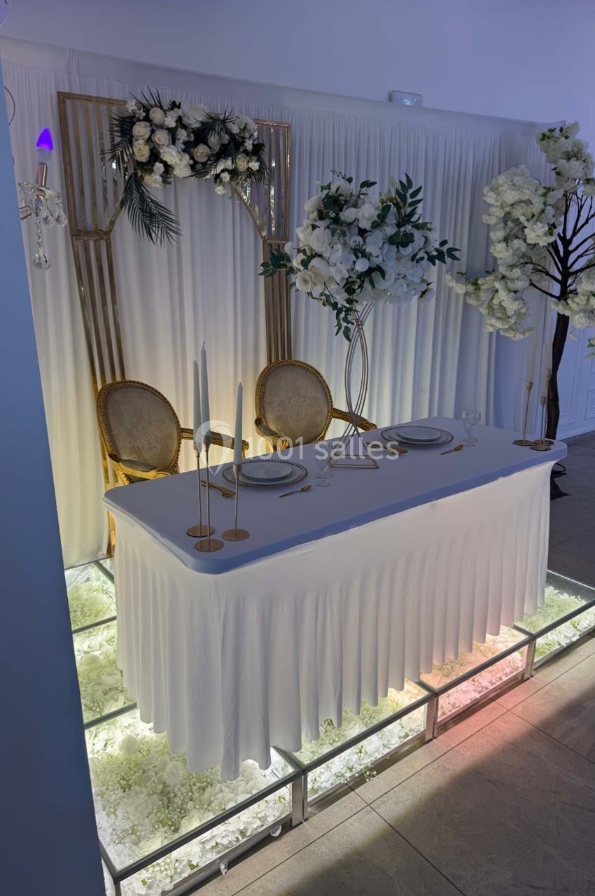 Table décorée pour un mariage avec nappes blanches, chandeliers, assiettes élégantes et fleurs blanches en arrière-plan.