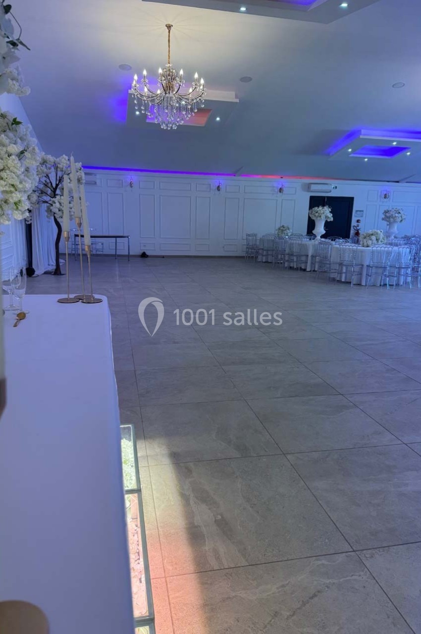 Salle de réception lumineuse avec sol en carrelage, tables décorées de fleurs blanches et lustre central élégant.