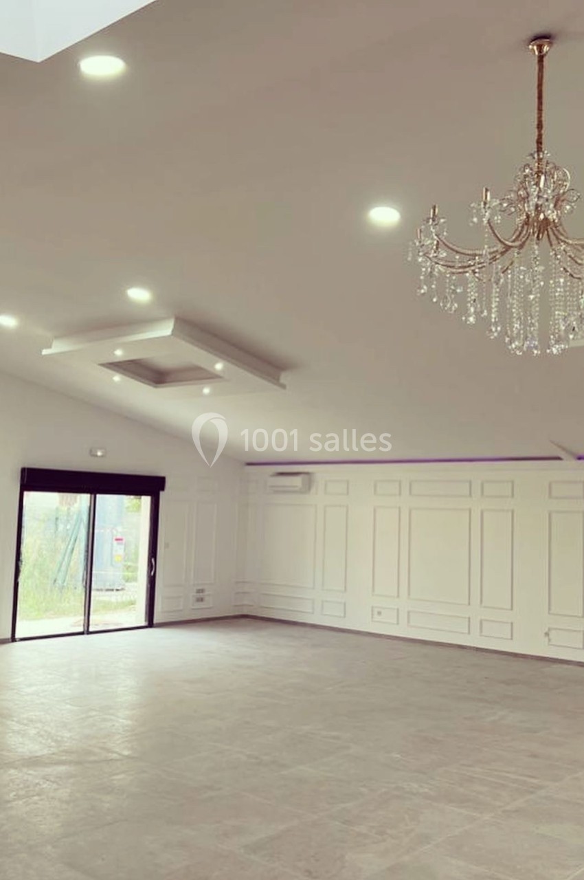 Salle vide avec sol en carrelage, murs blancs ornés de moulures, lustres suspendus et grande baie vitrée donnant sur l…