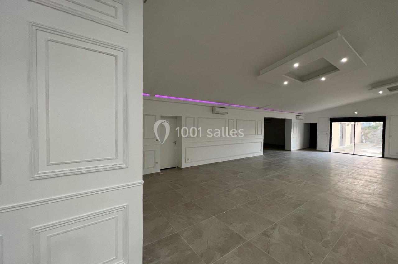 Salle spacieuse avec sol en carrelage, murs blancs moulurés, éclairage encastré et grande ouverture vitrée.