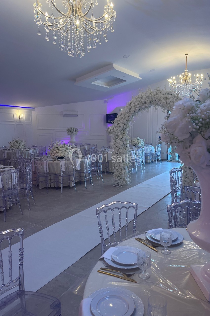 Salle de réception élégante avec tables dressées, chaises transparentes, arche florale blanche et lustres suspendus.