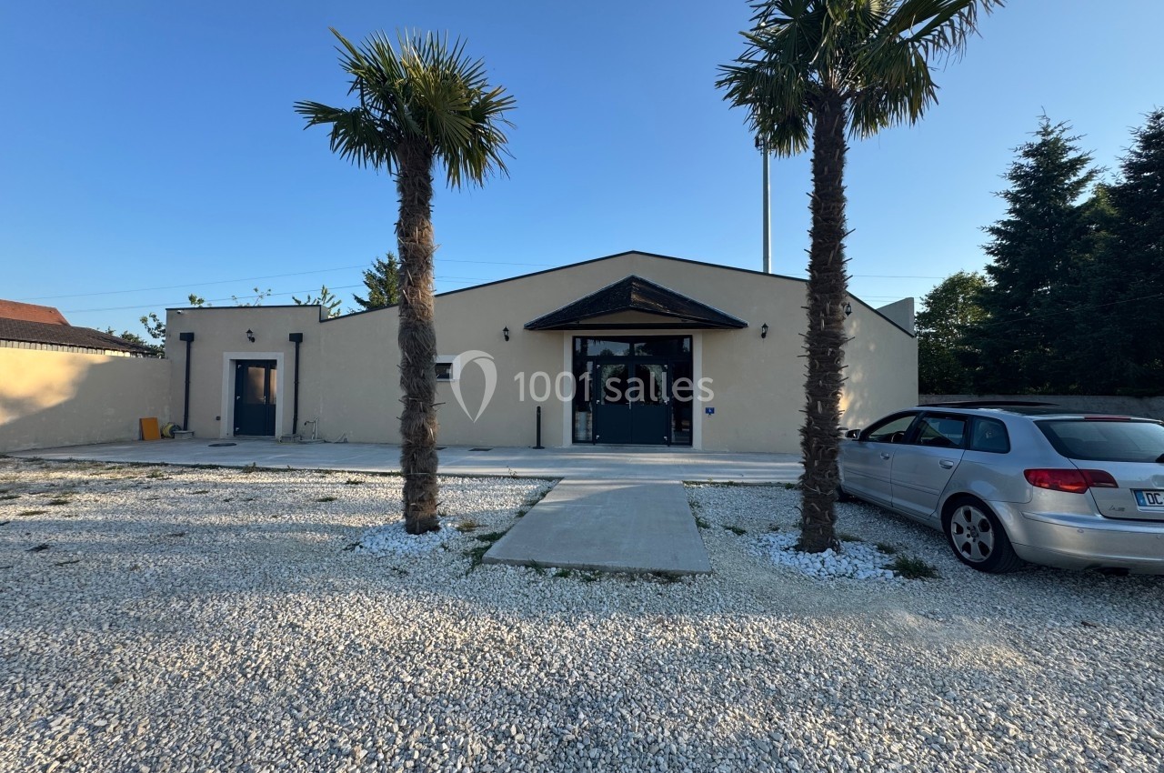 Façade d'un bâtiment beige avec deux palmiers devant, une voiture stationnée à droite et un sol en gravier.