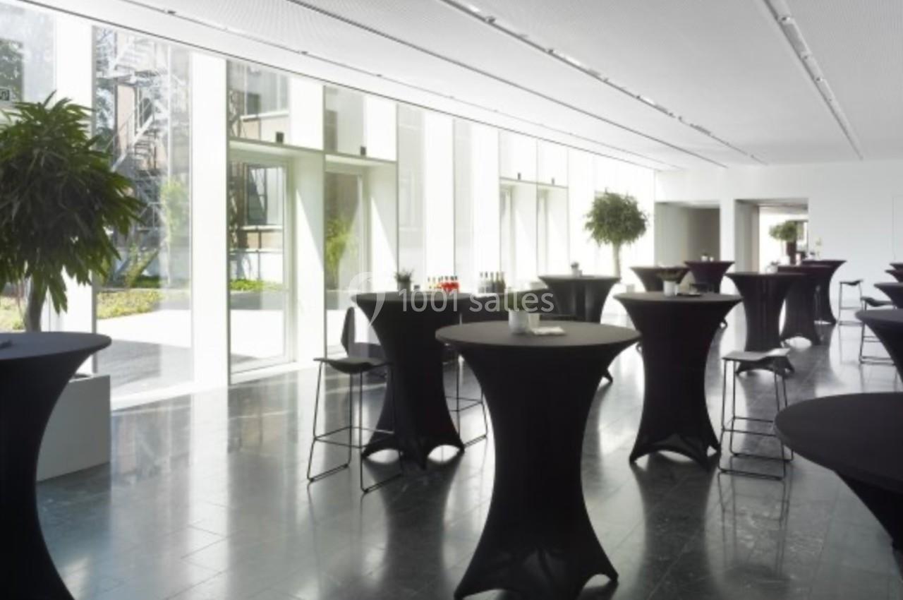 Salle lumineuse avec des tables hautes noires, des plantes en pot et des baies vitrées donnant sur un espace extérieur.