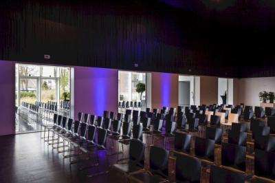 Salle de conférence vide avec rangées de chaises noires, éclairage bleu et une table haute sur une estrade.
