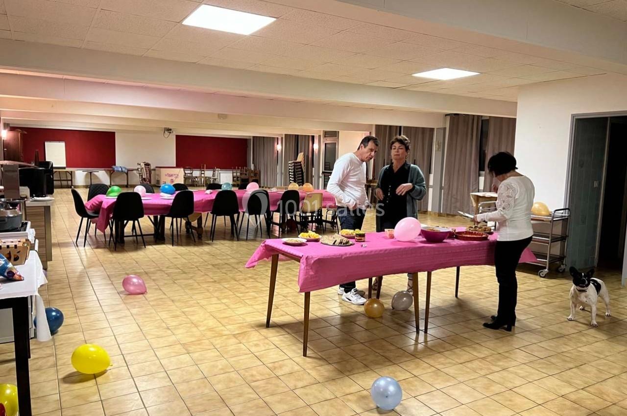 Salle décorée avec des ballons, trois personnes discutent près d'une table avec nappes roses et collations.