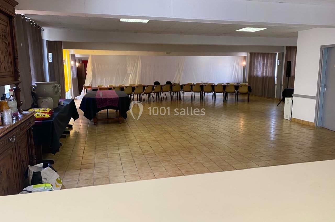 Salle vide avec sol carrelé, rangée de chaises alignées contre un mur blanc et tables recouvertes de nappes.