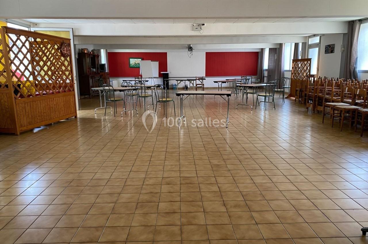 Salle vide avec tables et chaises empilées, sol carrelé, murs blancs et rouges, éclairée par des fenêtres.