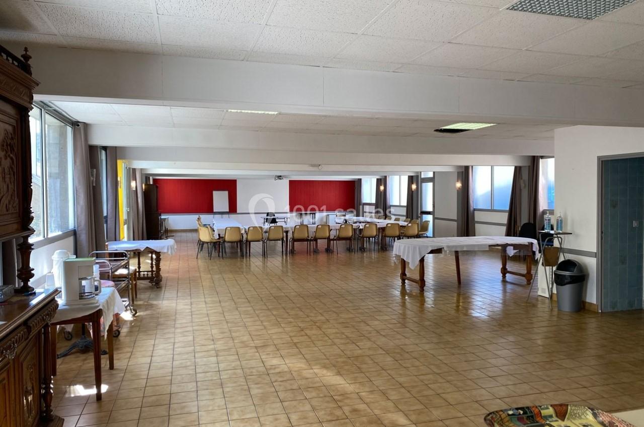 Salle spacieuse avec tables et chaises disposées, murs rouges en arrière-plan et lumière naturelle entrant par les fenêtres.