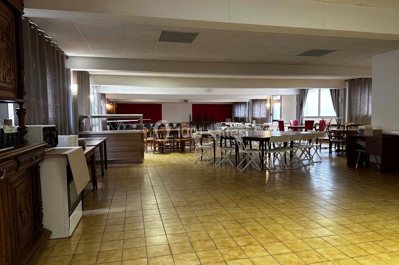 Salle spacieuse avec tables et chaises disposées pour un événement, sol en carrelage jaune et rideaux gris aux fenêtres.