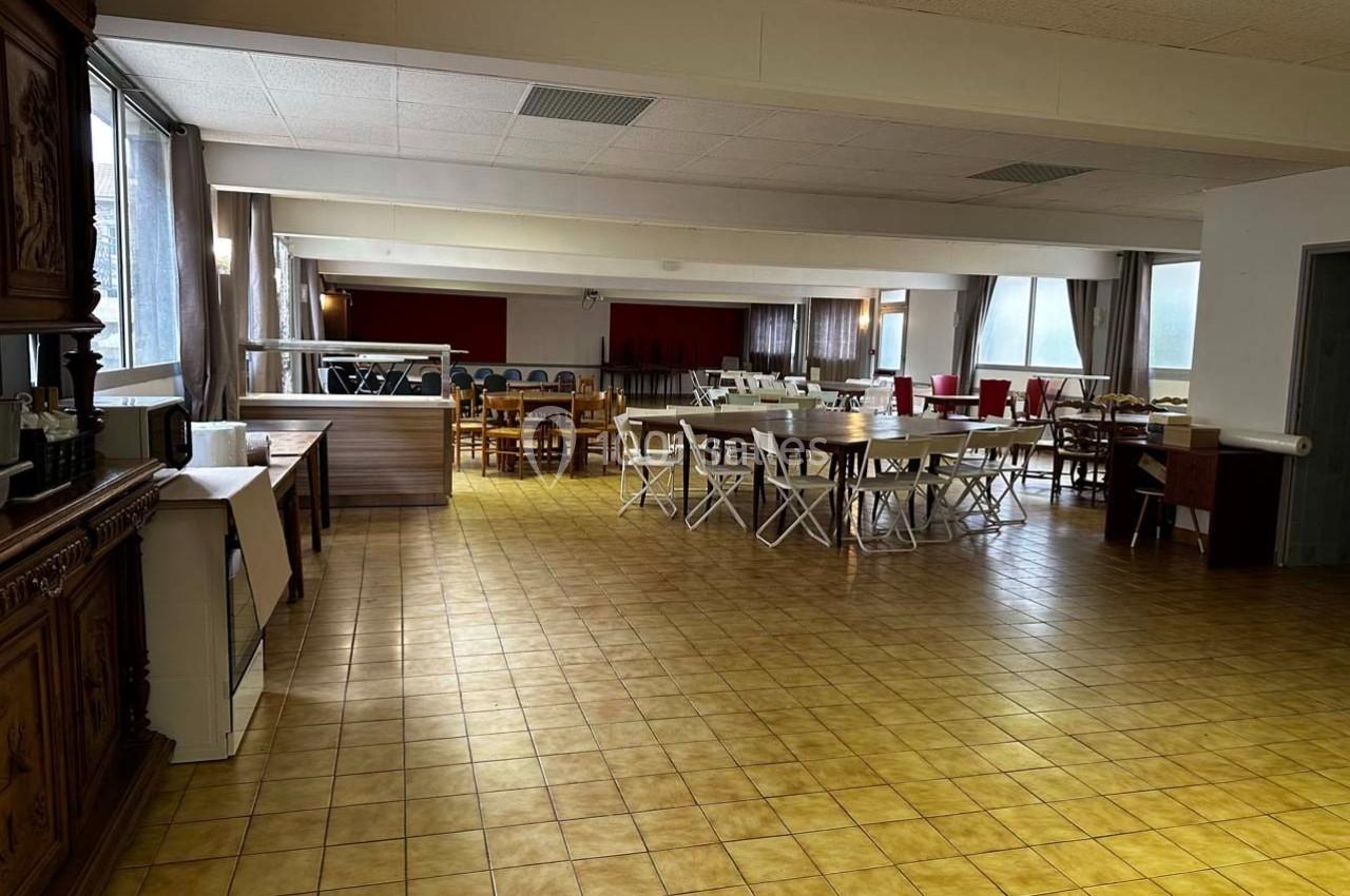 Salle spacieuse avec tables et chaises disposées pour un événement, éclairée par de grandes fenêtres.